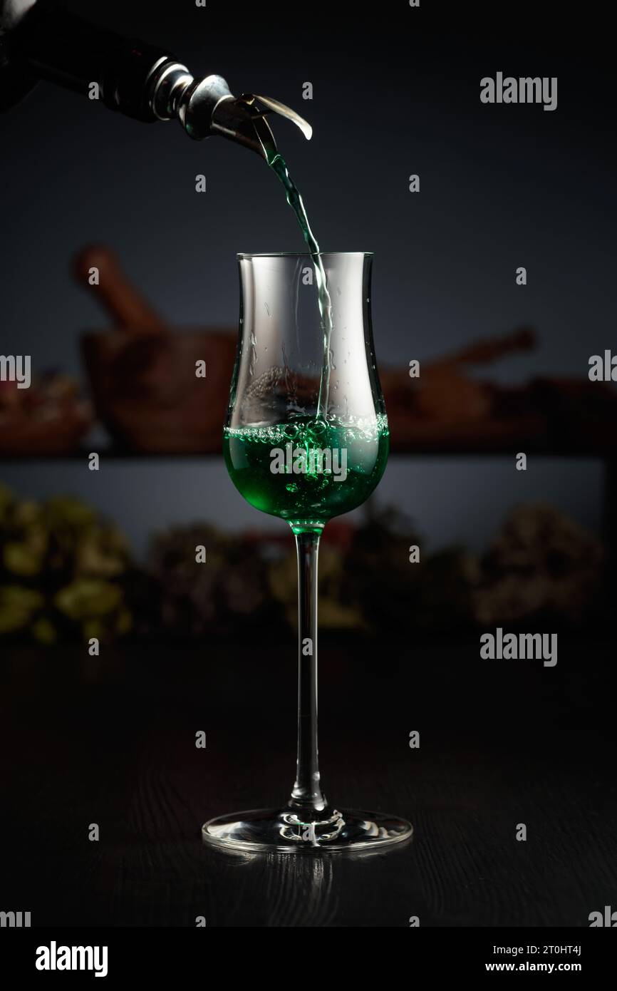 Grüner Kräuterlikör wird aus einer Vintage-Flasche in ein Glas gegossen. Im Hintergrund getrocknete Kräuter und alte Küchenutensilien. Stockfoto