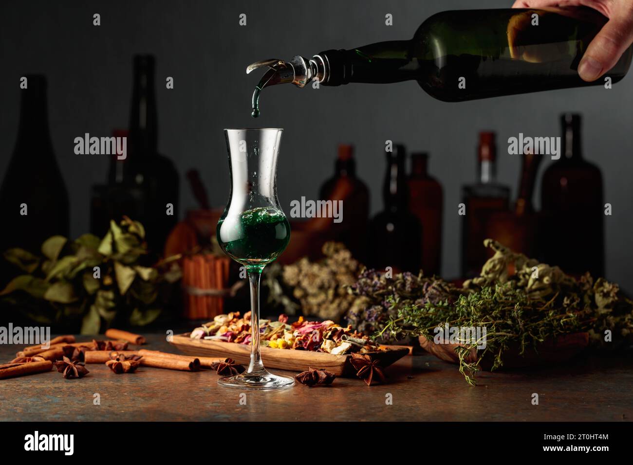 Grüner Kräuterlikör wird aus einer Vintage-Flasche in ein Glas gegossen. Auf einem Tisch getrocknete Kräuter, Blumen, Gewürze und alte Küchenutensilien. Kräuterbegriff Stockfoto