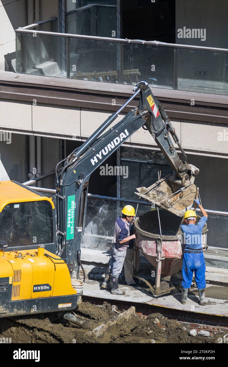 An einem sonnigen Tag entlädt eine Gruppe von Bauarbeitern vorgemischten Zement, der von einem Gabelstapler in der Nähe des im Bau befindlichen Gebäudes gebracht wird. Stockfoto