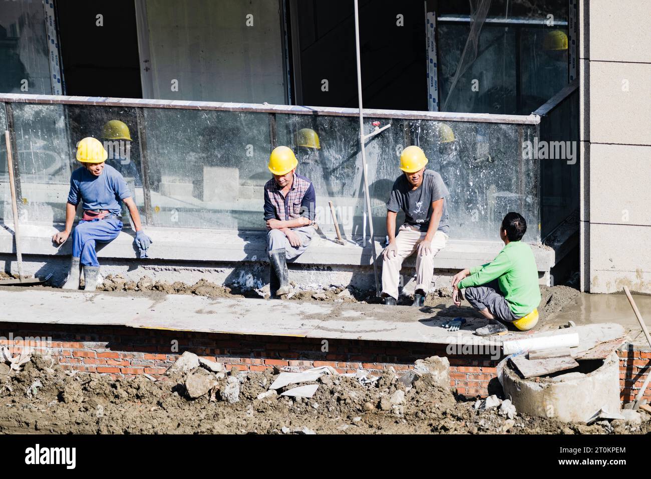 An einem sonnigen Tag sitzen einige Arbeiter an der Mauer, um sich auszuruhen, während andere auf ihren Hüten sitzen, um eine Pause einzulegen. Stockfoto