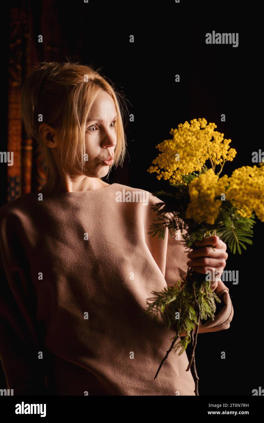 In einem dunklen Raum sitzt eine anmutige Frau mittleren Alters mit einem Haufen gelber Blumen, ihr Gesicht reflektiert schieres Staunen Stockfoto