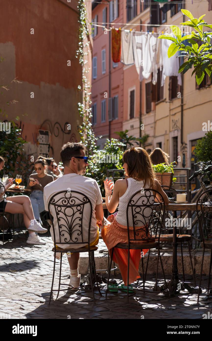 Ein junges Paar, das sich gerne unterhalten kann, sitzt vor einer Bar in einer typischen engen Gasse in Trastevere, dem historischen Viertel von Rom, Italien, Europa, EU Stockfoto