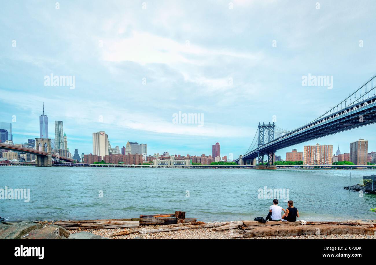 Ein junges Paar sitzt am Fluss und genießt die Skyline von Lower Manhattan. Die Brooklyn Bridge befindet sich auf der linken Seite und die Manhattan Bridge auf der rechten Seite. Stockfoto