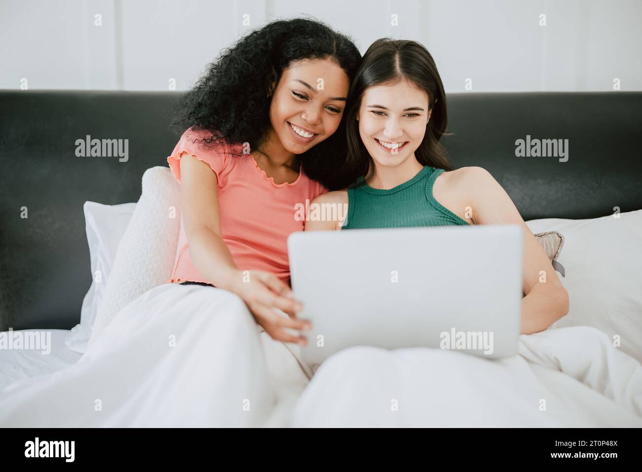 LGBT-Frauen, die auf dem Bett sitzen und auf dem Laptop-Bildschirm sehen, glücklich lächelnd, zusammen zu Hause Urlaub Aktivität Stockfoto