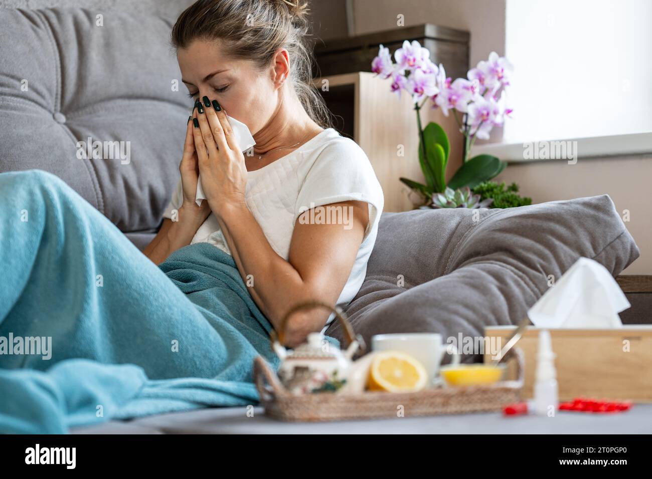 Frau niest in ein Taschentuch. Eine kranke Frau, die zu Hause unter einer Decke sitzt. Vor ihr steht ein Tablett mit einer Teekanne, Zitrone und Honig. Stockfoto