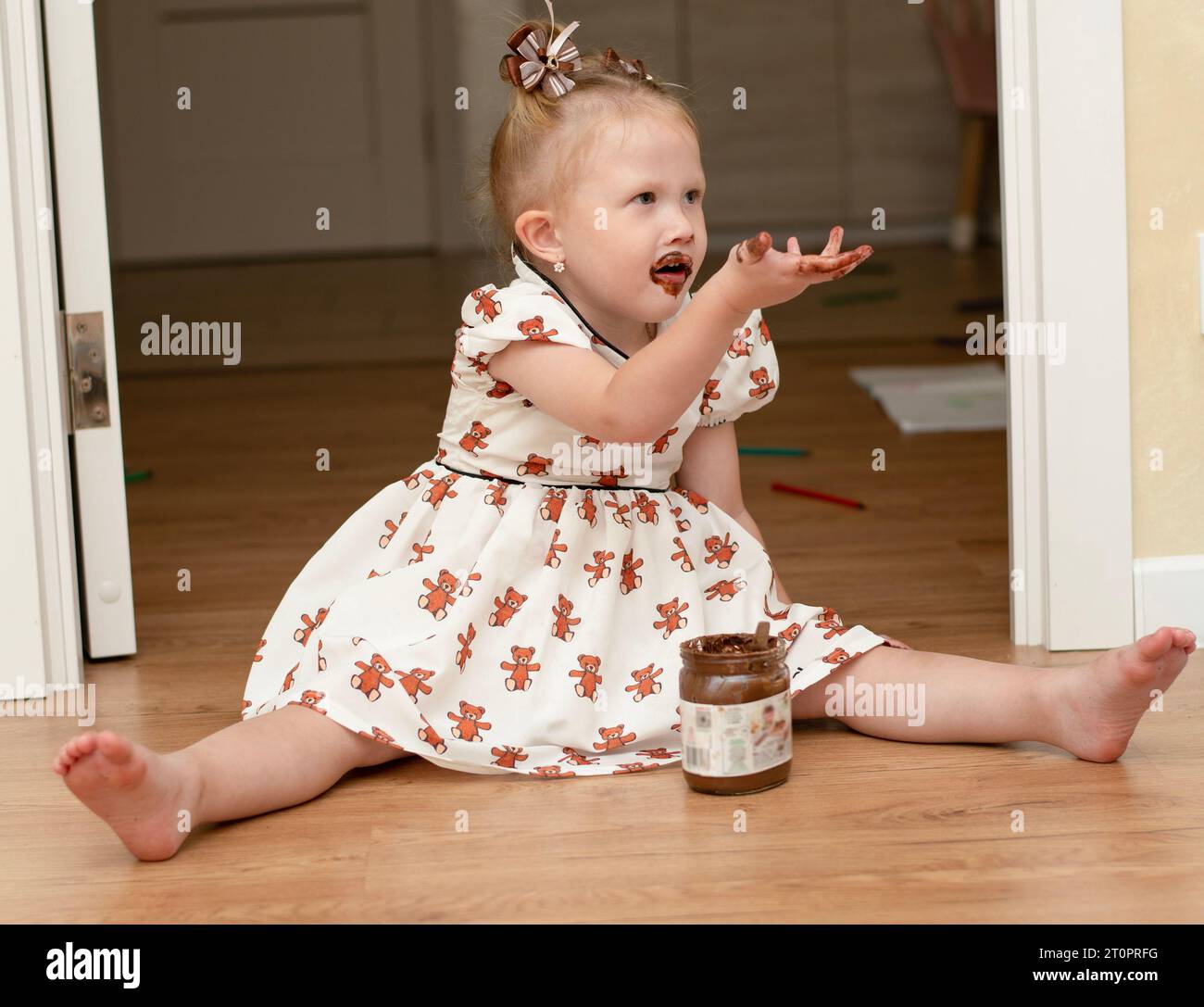 Ein kleines Mädchen, 3 Jahre alt, in einem schönen weißen Kleid, sitzt auf dem Boden in einem Wohnraum und isst glücklich Schokolade aus einem Glas. Sein gesamter Mout Stockfoto