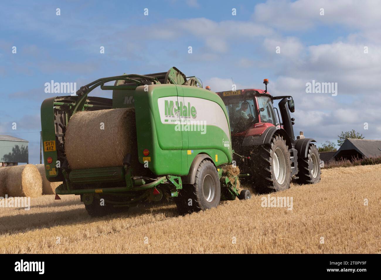 Die Rückseite einer McHale-Ballenpresse und eines Red Valtra-Traktors, die am Ende der Ernte auf einem Stoppelfeld Stoppelballen herstellen Stockfoto
