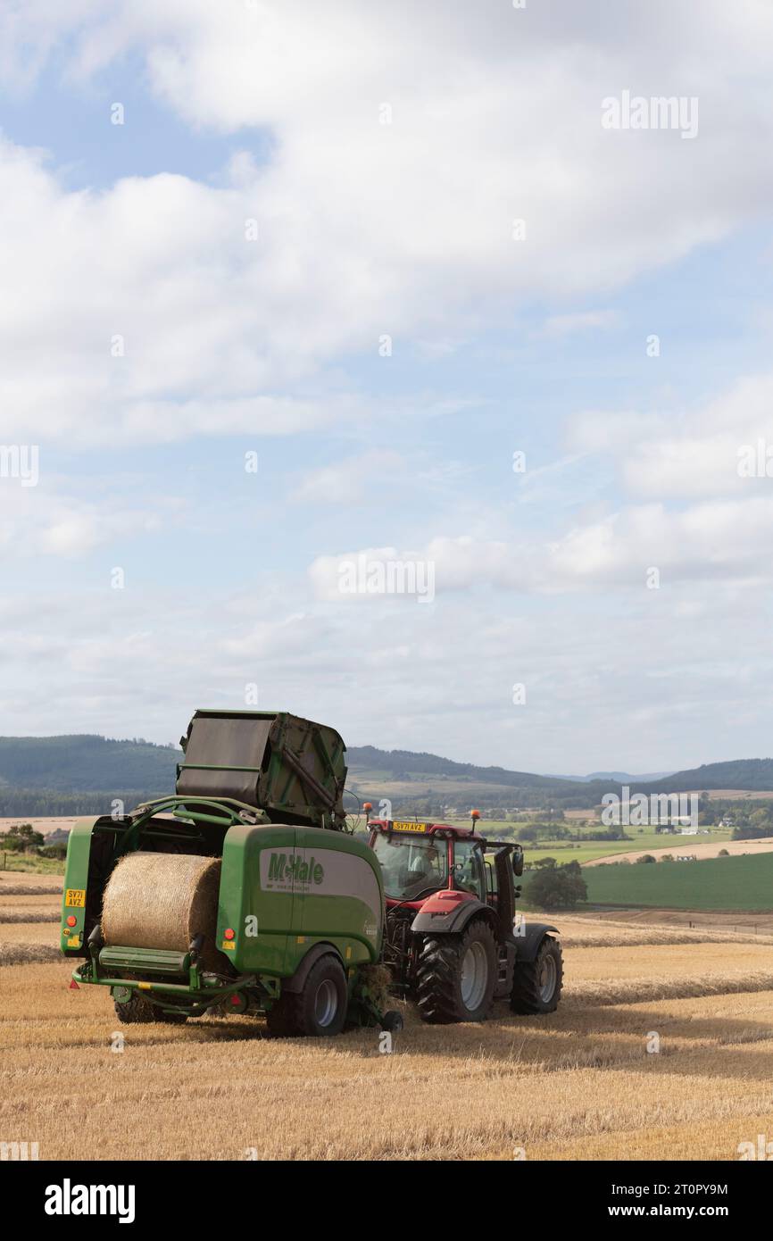 Rückansicht einer McHale-Ballenpresse, die von einem Red Valtra-Traktor gezogen wird und einen Strohballen produziert, mit Blick auf die umliegende Landschaft von Aberdeenshire Stockfoto