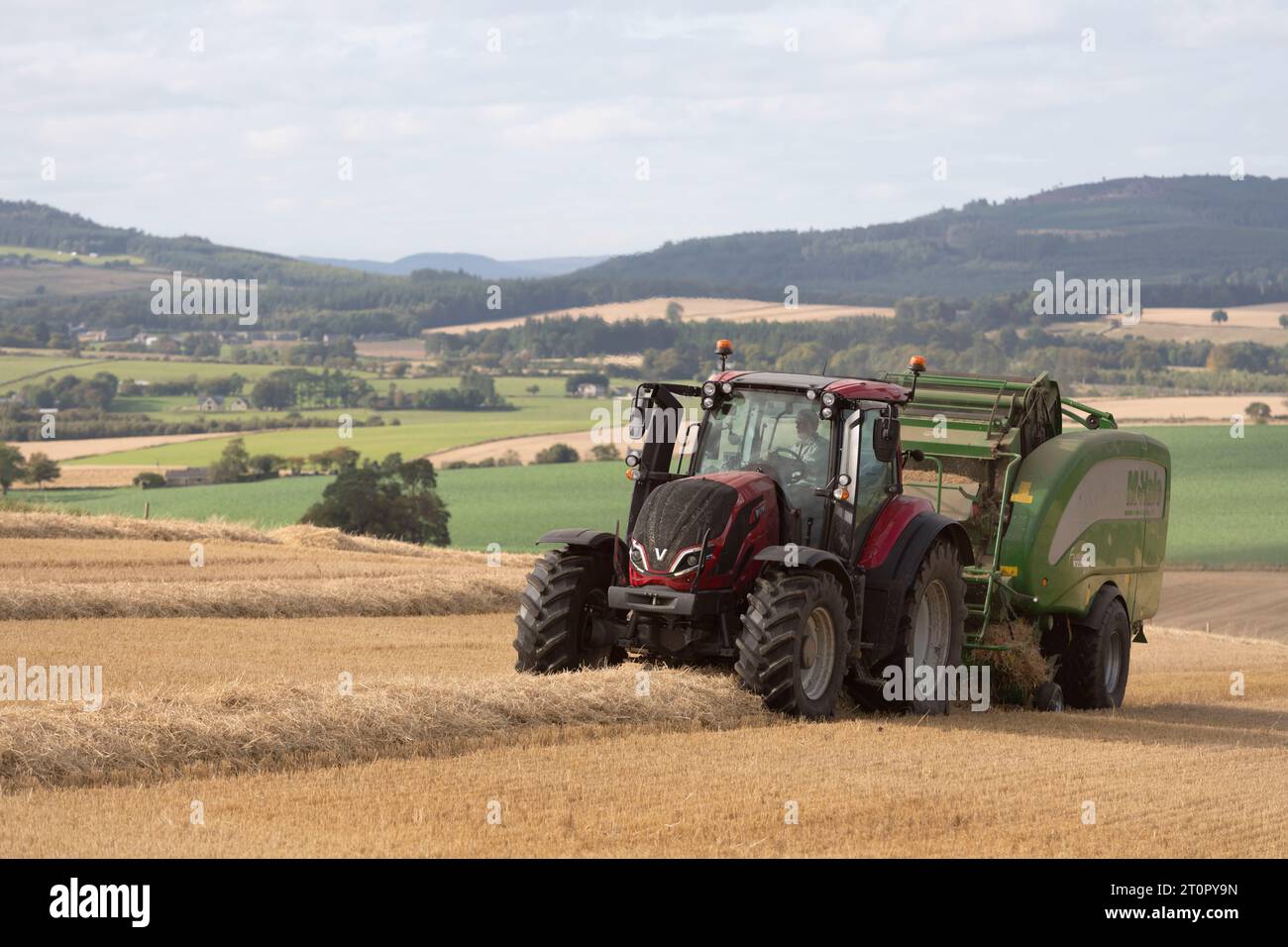 Ein Red Valtra-Traktor mit einer McHale-Ballenpresse, der auf einem Stoppelfeld arbeitet und einen Blick über die malerische Landschaft von Aberdeenshire bietet Stockfoto