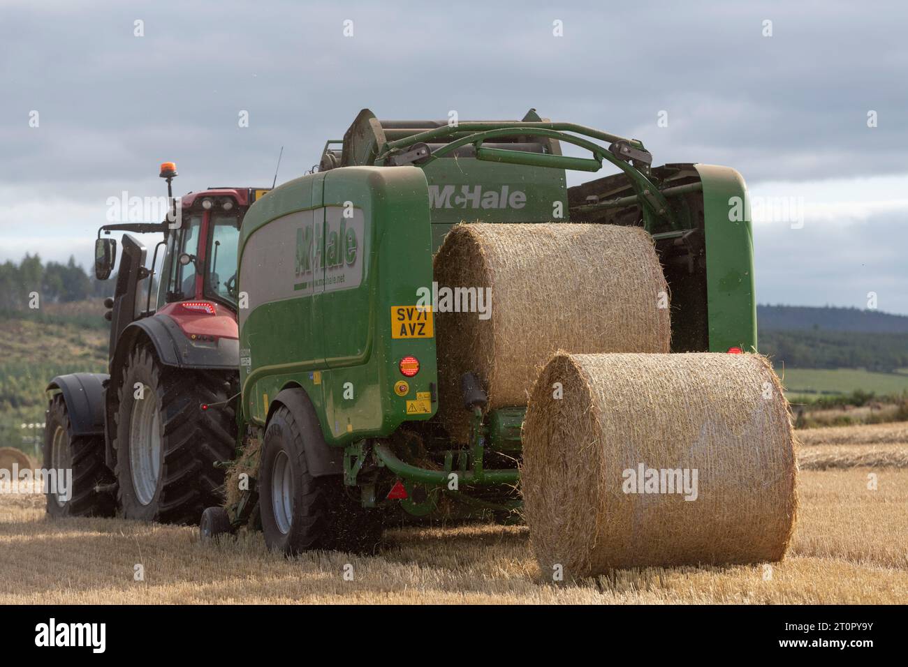 Eine McHale-Ballenpresse, die von einem Red Valtra-Traktor gezogen wird und während der Ernte von hinten gesehen zwei Strohballen auf einem Stoppelfeld platziert Stockfoto