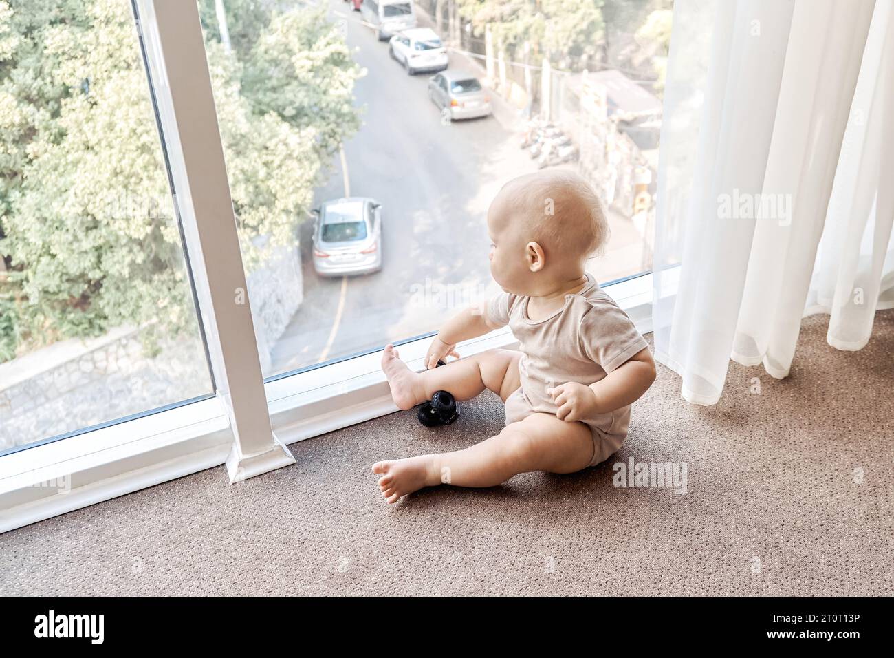 Neugieriges Baby schaut aus dem Panoramafenster, das auf dem Boden sitzt. Niedliches Kleinkind liegt in der Nähe eines großen Fensters im Hotelzimmer. Gesundes Kind zu Hause Stockfoto