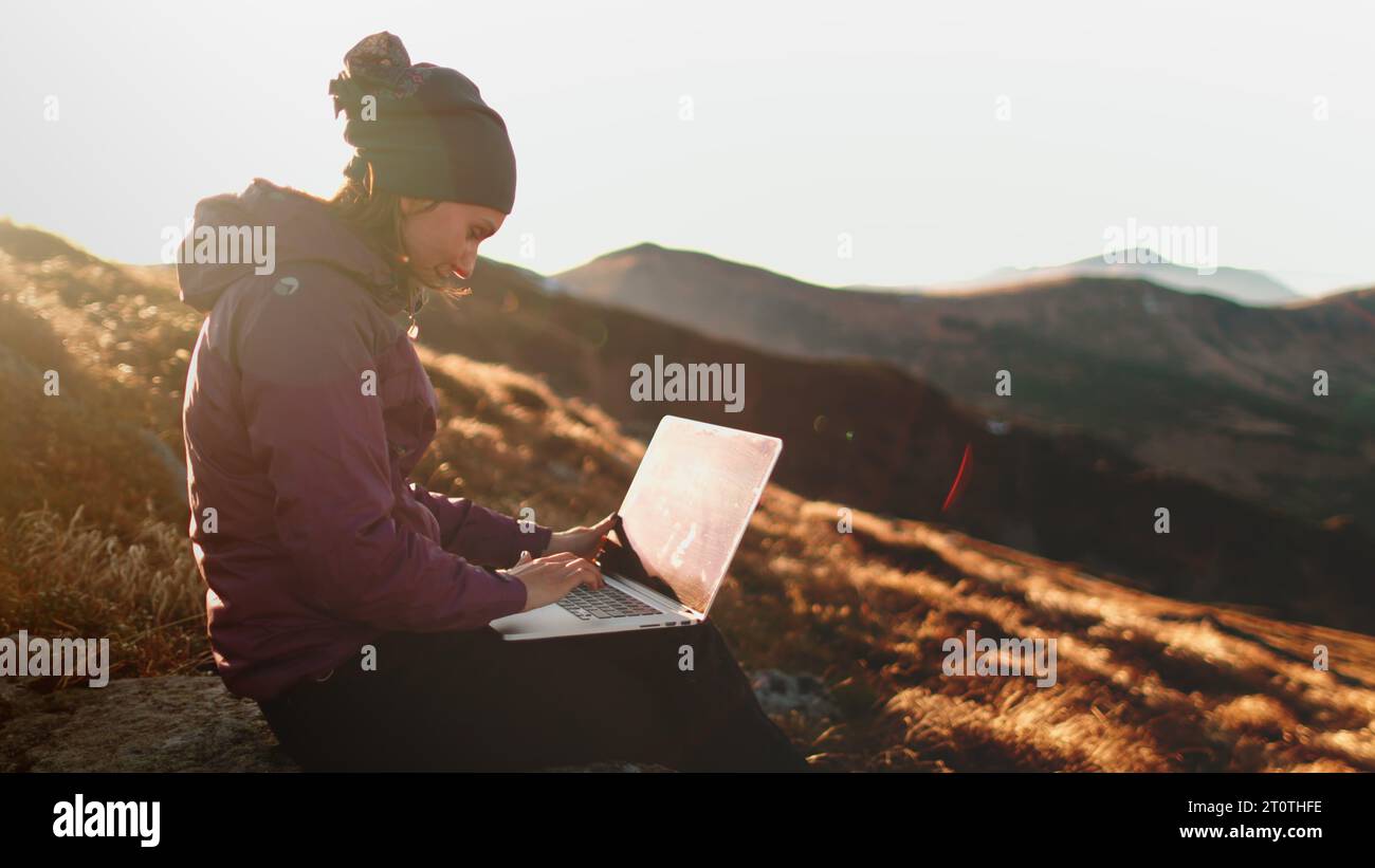 Herbst Sonnenuntergang Bergfrau, die per Fernbedienung am Laptop arbeitet. Touristenmädchen, das auf einem Notizbuch sitzt, sitzt auf einer orangefarbenen Graswiese in sanftem Sonnenlicht. Wandern, Trekking Stockfoto