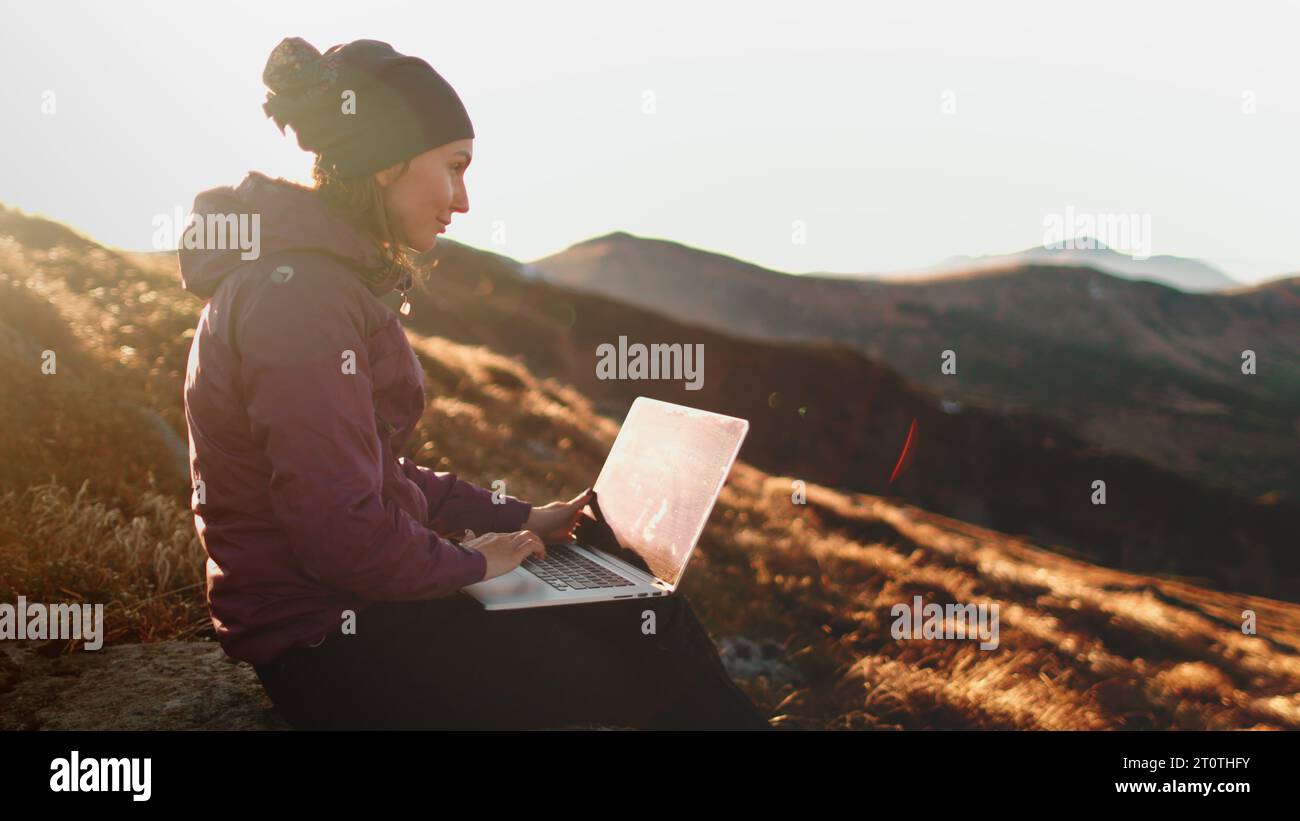 Herbst Sonnenuntergang Bergfrau, die per Fernbedienung am Laptop arbeitet. Touristenmädchen, das auf einem Notizbuch sitzt, sitzt auf einer orangefarbenen Graswiese in sanftem Sonnenlicht. Wandern, Trekking, Outdoor Lifestyle Reisen im Urlaub Stockfoto