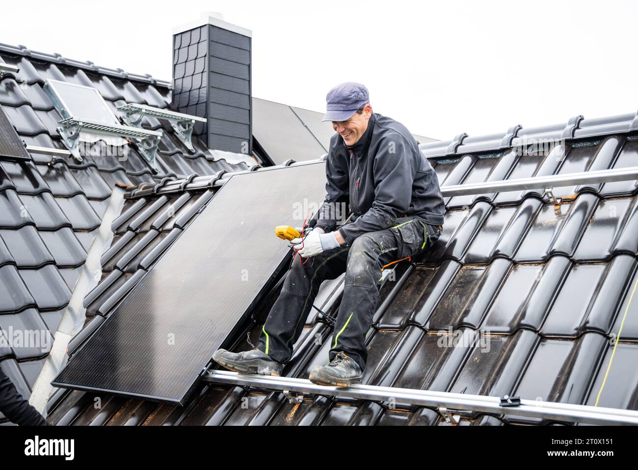 Ein lächelnder Techniker sitzt auf dem Dach eines Hauses neben einem Sonnenkollektor Stockfoto