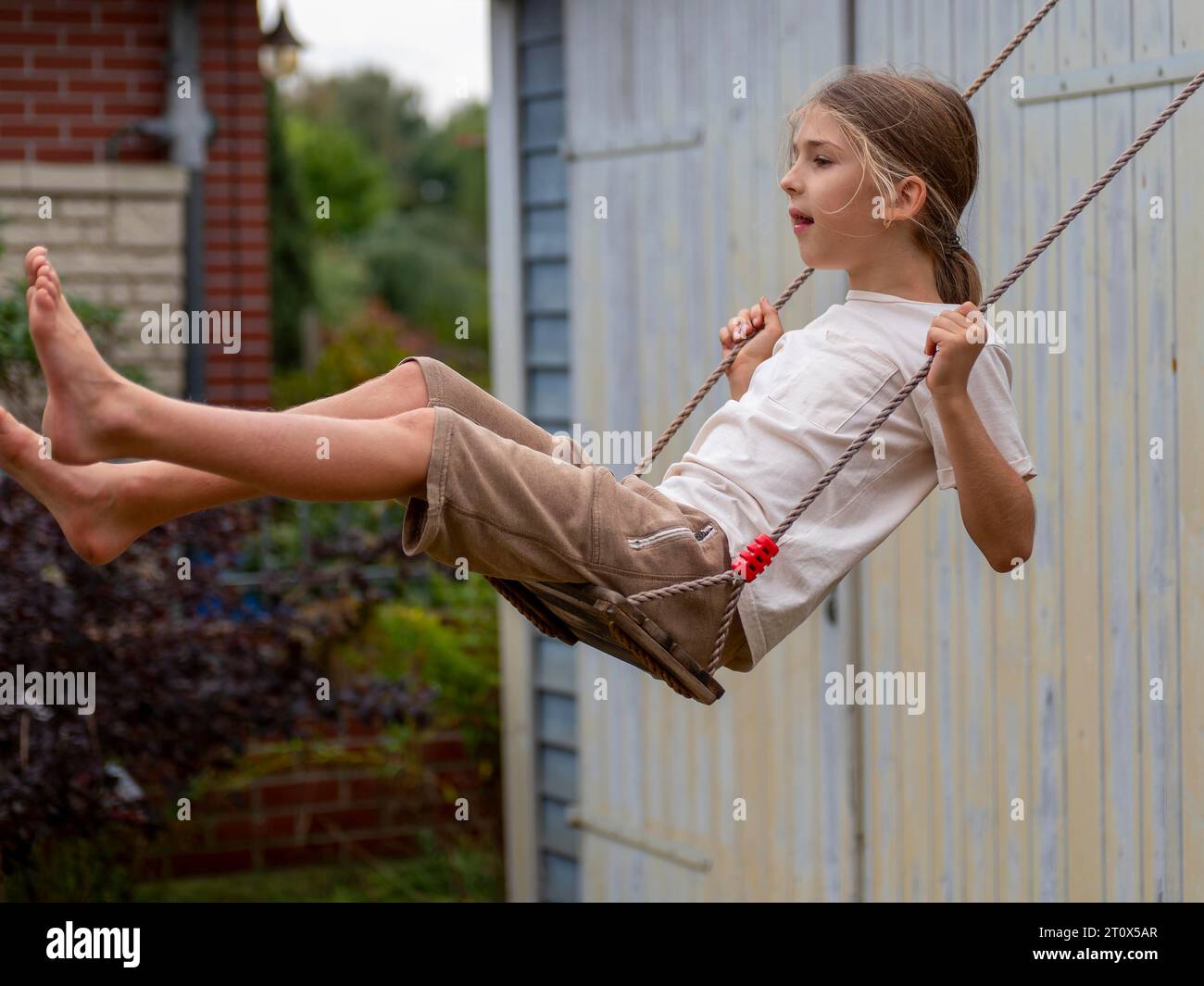 Junge schwingend, während er auf der Kinderschaukel am Schuppen und Gartentor sitzt, Deutschland Stockfoto