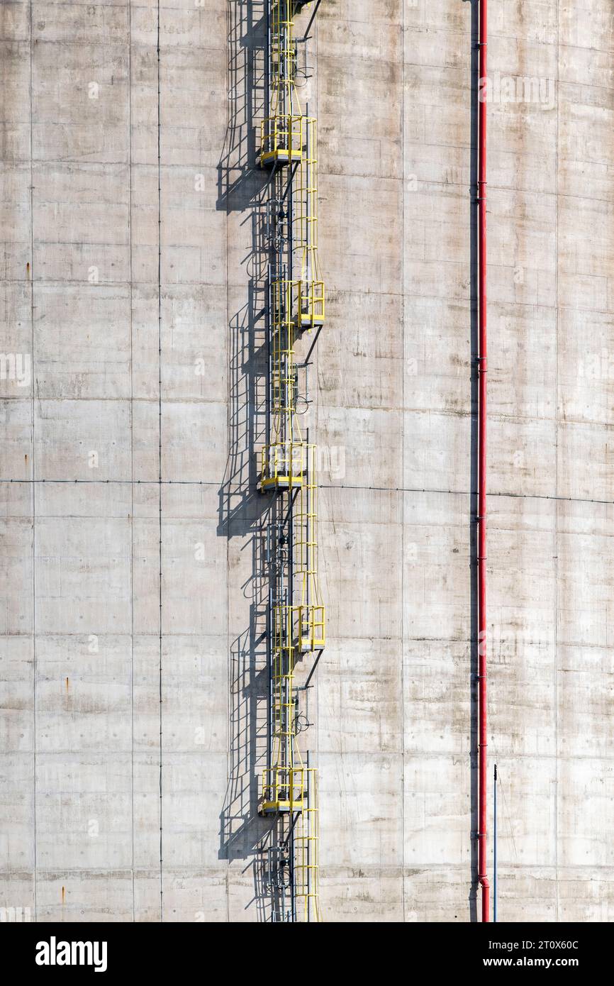 Bildfüllungsansicht von Sichtbeton an der Seite eines Industrietanks mit gelber vertikaler Leiter mit Sicherheitskäfig und roter vertikaler Rohrleitung für W Stockfoto