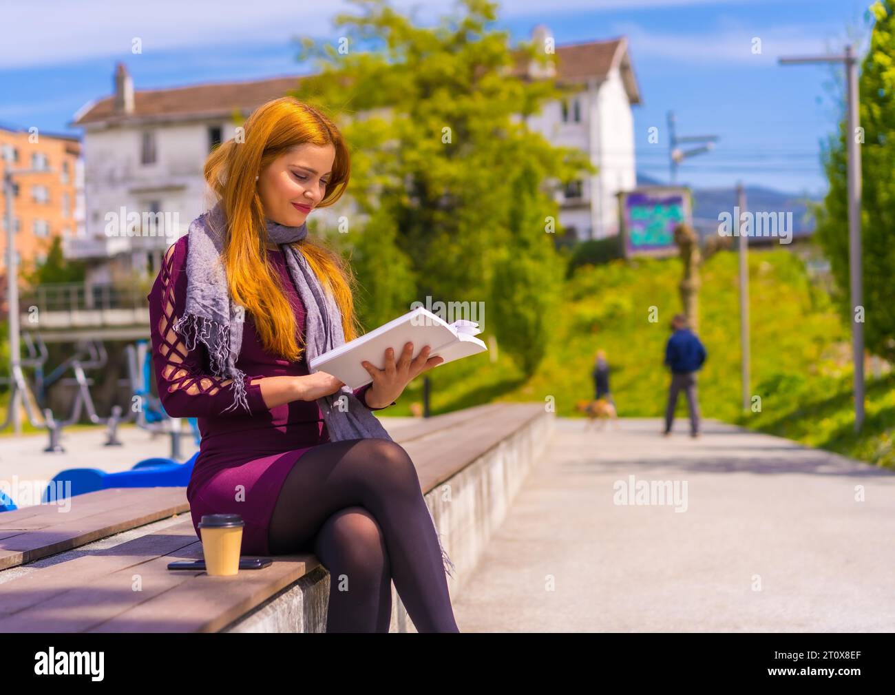 Schöne Frau in lila Kleid und Schal liest ein Buch in einem Park in der Stadt Stockfoto