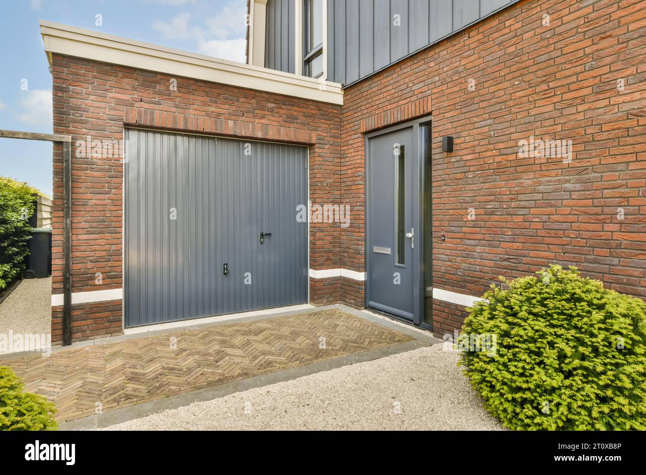 Ein graues Garagentor vor einer roten Backsteinwand mit grünen Büschen und Sträuchern auf beiden Seiten des Gebäudes Stockfoto