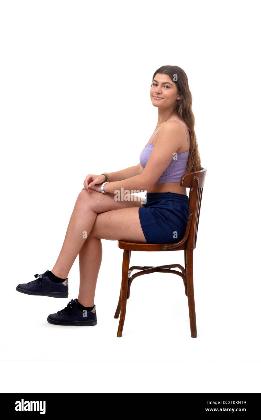 Seitenansicht eines jungen Mädchens, das auf Stuhl sitzt und in die Kamera blickt und mit den Beinen auf weißem Hintergrund Stockfoto