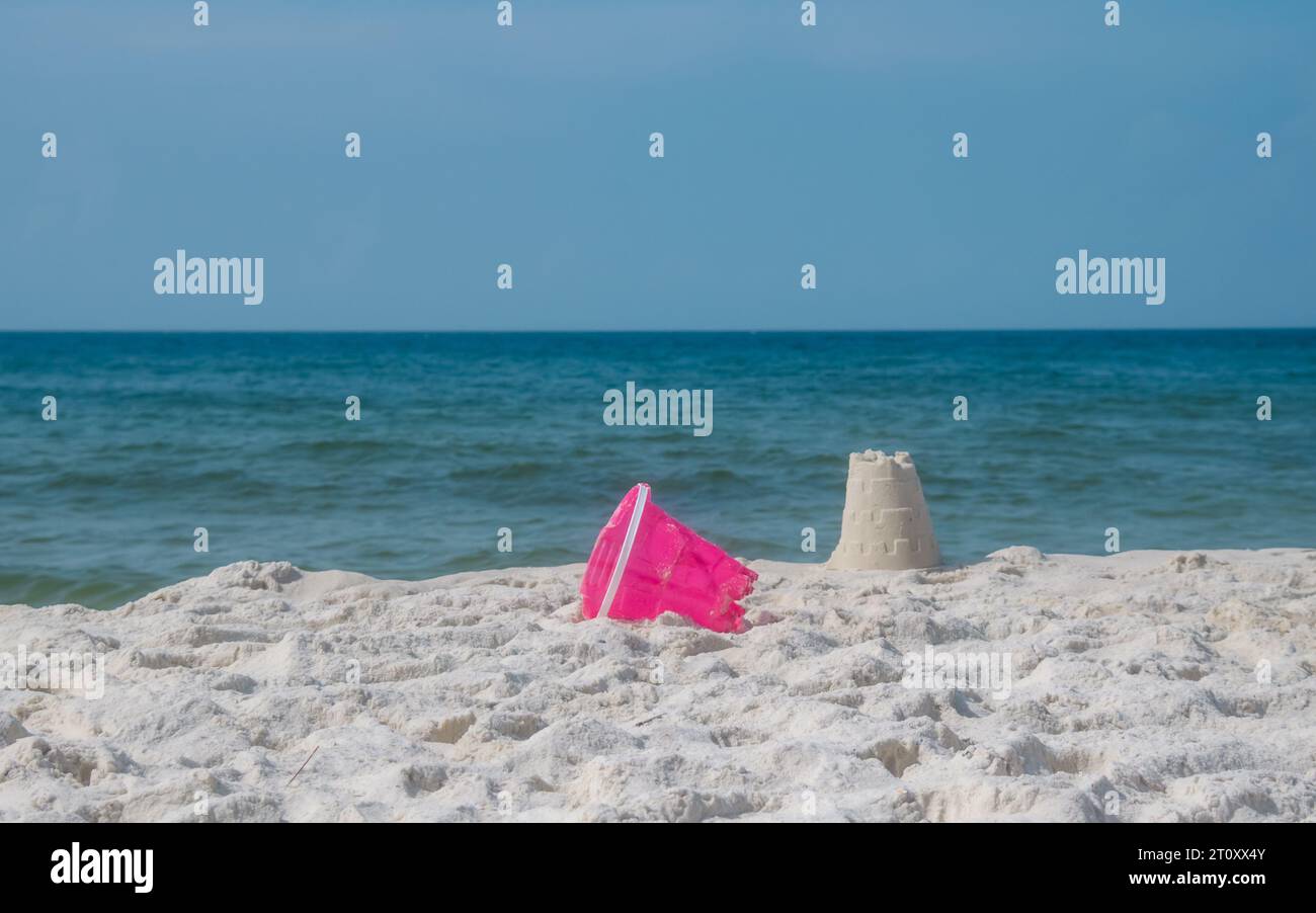 Sandburg auf weißem Meer Strand Sand. Farbenfroher rosafarbener Spielzeugeimer, der neben dem Sandschloss am Meer liegt. Ein unterhaltsamer Tag voller Freizeit und Spiel am Strand. Stockfoto