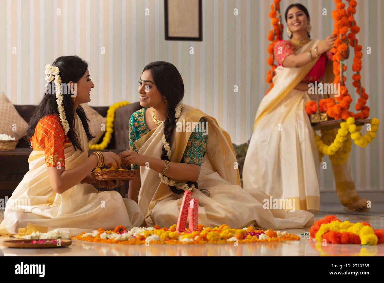 Zwei Frauen dekorieren den Boden mit Blumen auf Onam, während andere Frauen auf einer Schaukel sitzen Stockfoto