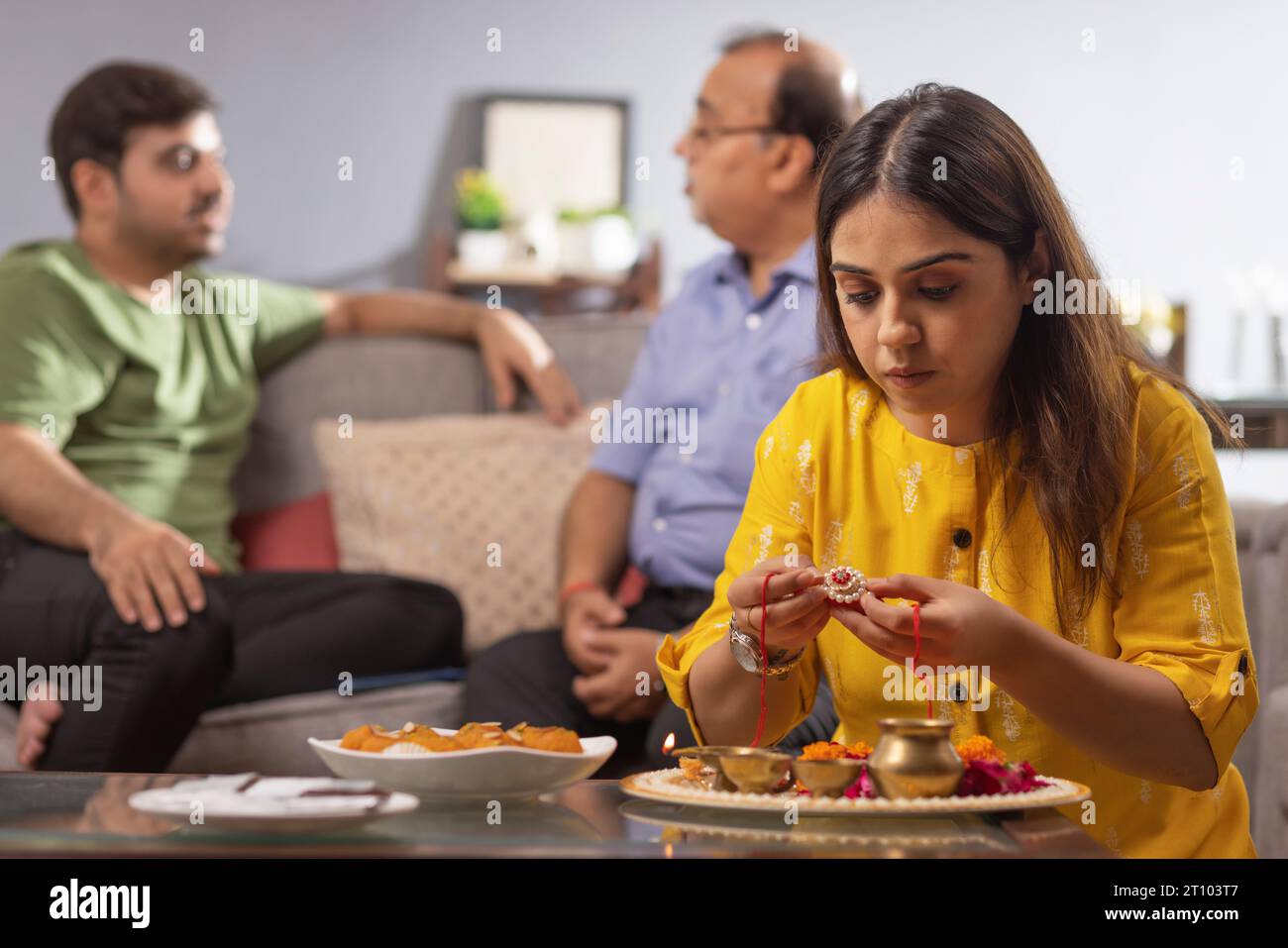 Eine junge Frau arrangiert Puja Thali anlässlich von Raksha Bandhan, während ihr Vater und Bruder dahinter sitzen Stockfoto