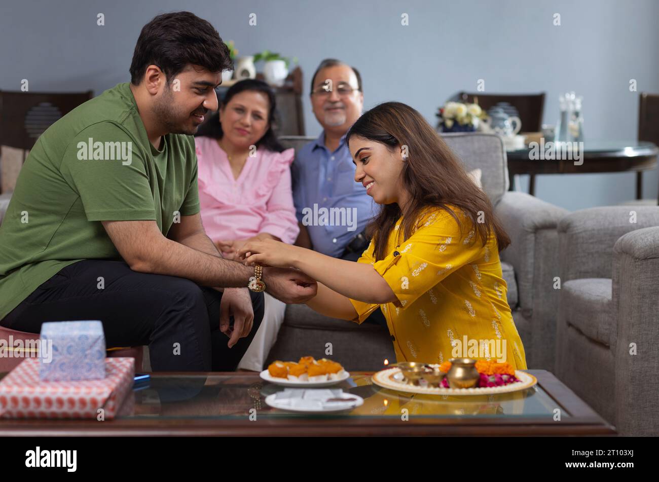 Schwester fesselt Rakhi am Handgelenk ihres Bruders an Raksha Bandhan, während die Eltern nebenan sitzen Stockfoto