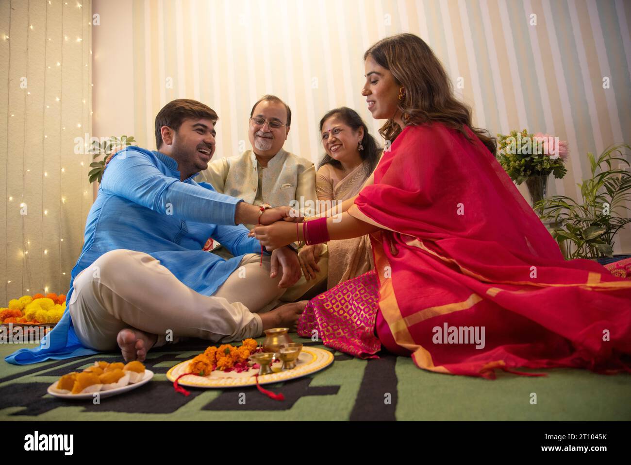 Schwester fesselt Rakhi am Handgelenk ihres Bruders an Raksha Bandhan, während die Eltern nebenan sitzen Stockfoto