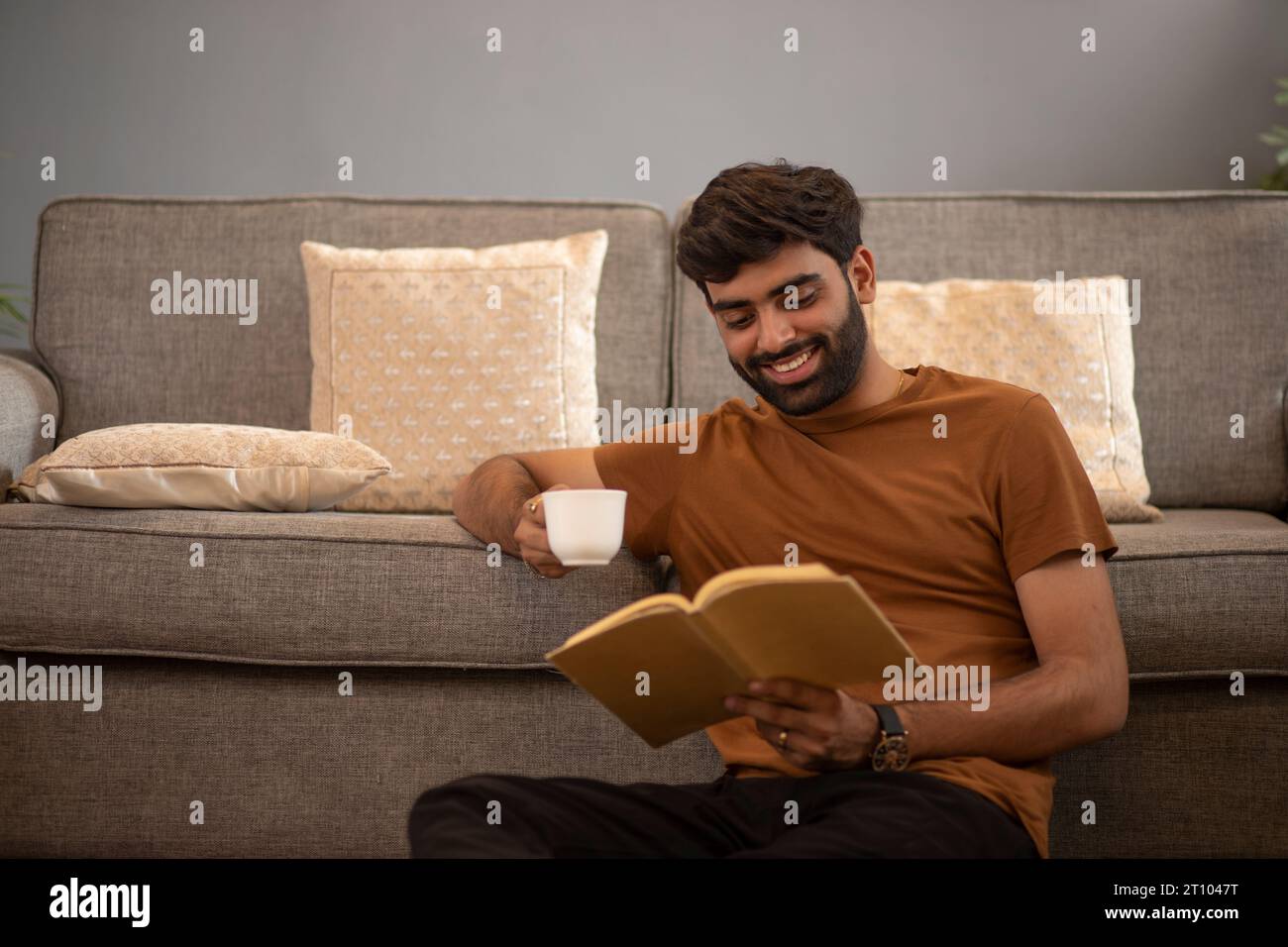 Ein lächelnder junger Mann trinkt Tee, während er sich auf dem Sofa im Wohnzimmer entspannt Stockfoto