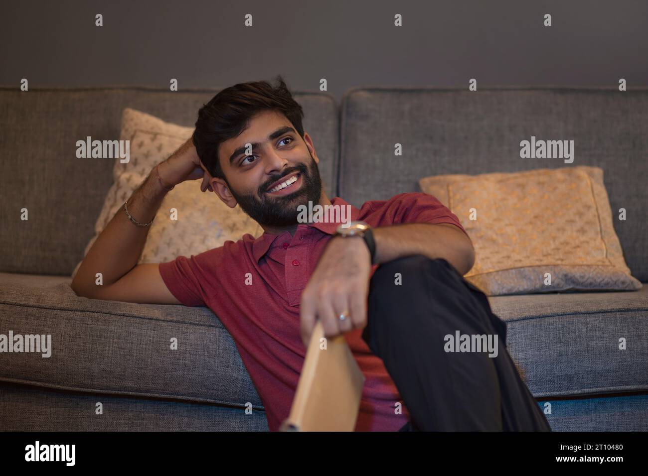 Ein junger Mann liest ein Buch, während er im Wohnzimmer auf dem Sofa liegt Stockfoto