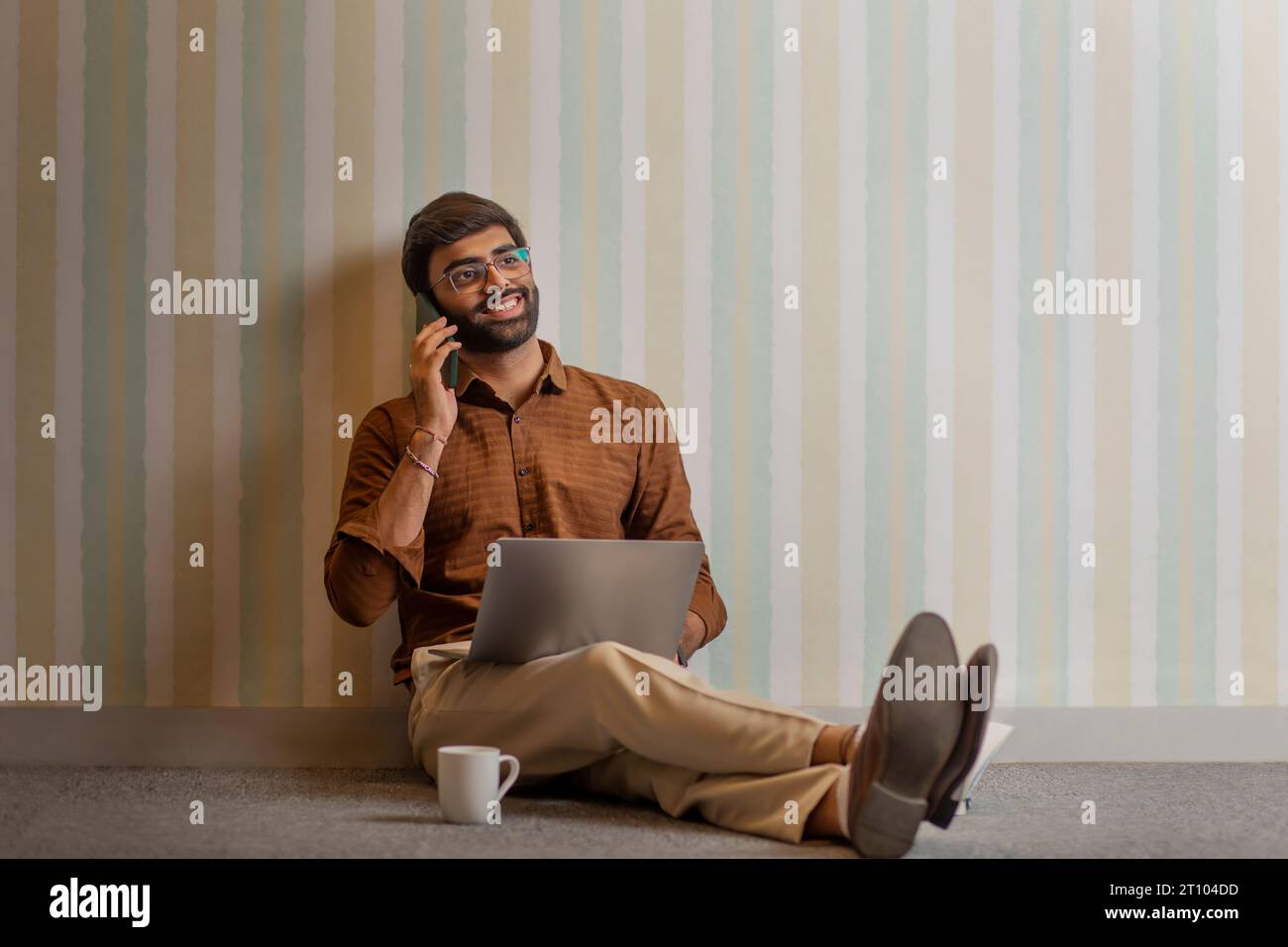 Junger Mann, der mit dem Handy spricht, während er in seinem Heimbüro mit einem Laptop arbeitet Stockfoto