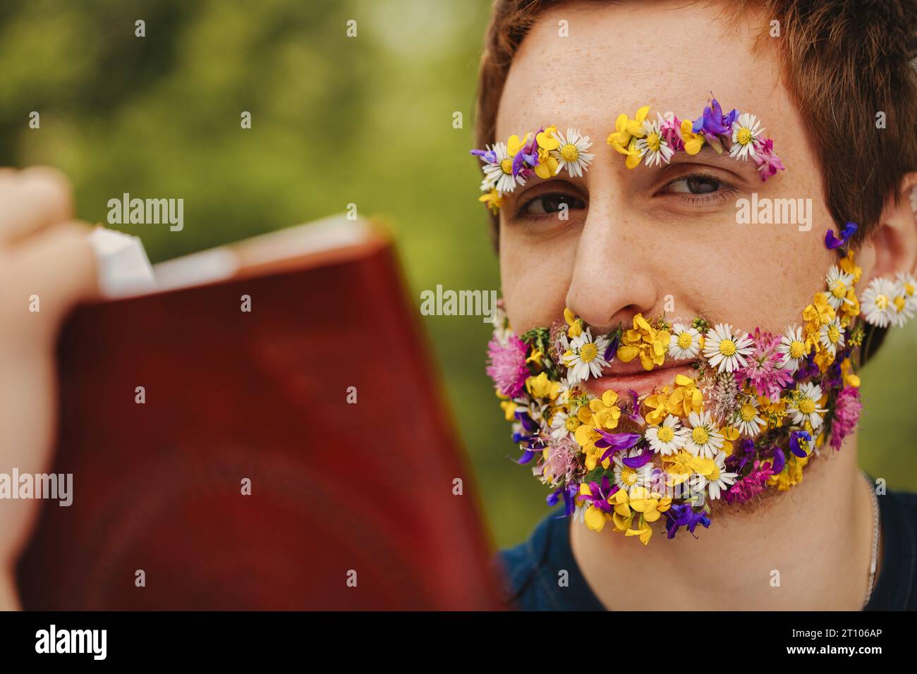 Tief im Wald zeigt ein junger Mann Frühlingsblumen anstelle von Gesichtshaaren, liest ein altes ledergebundenes Buch, das mit der Natur und dem li in Einklang steht Stockfoto