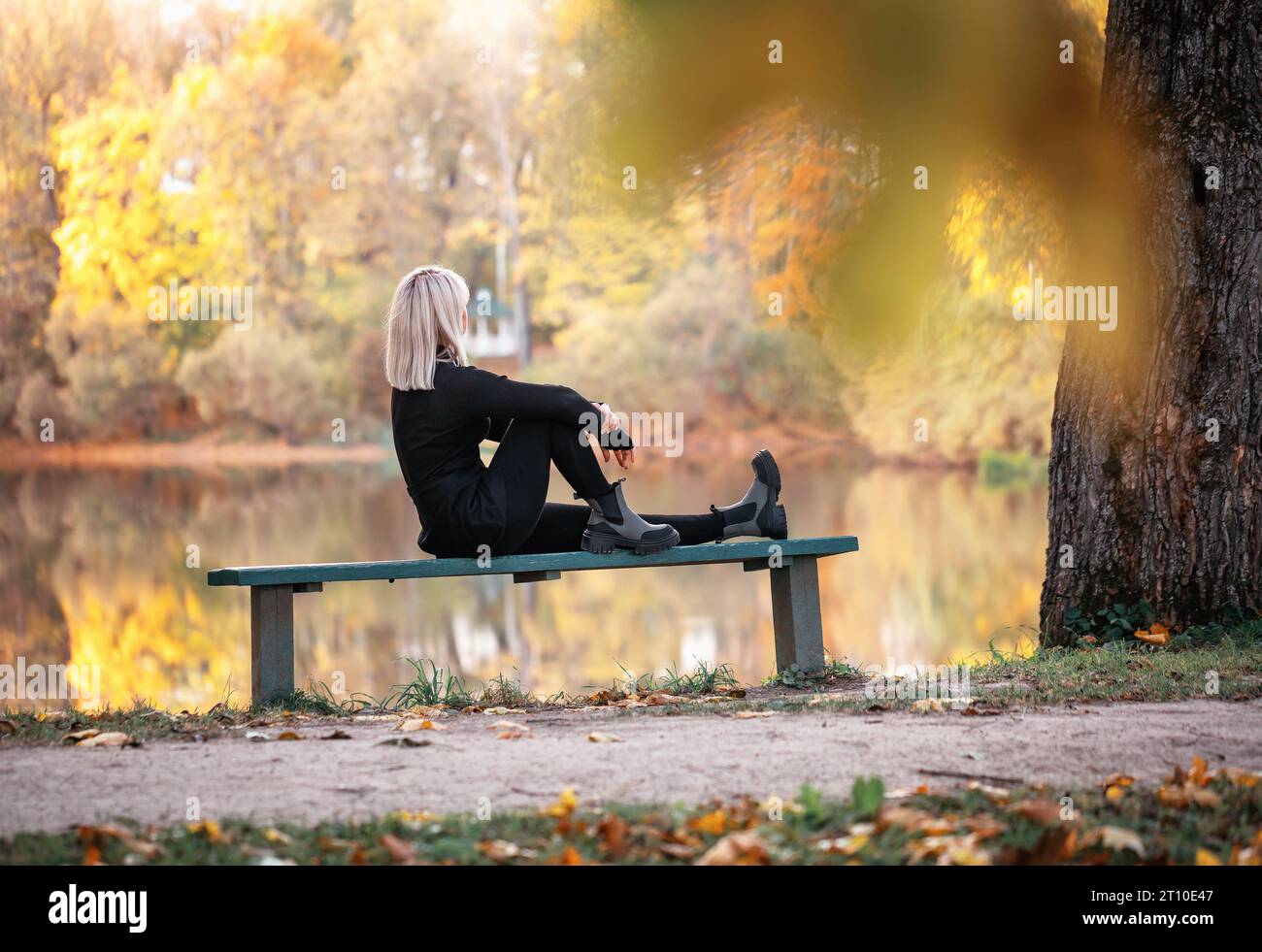 Eine blonde Frau entspannt sich an einem sonnigen Abend, sitzt auf einer Parkbank in der Nähe eines Teichs und bewundert den Herbstblick Stockfoto