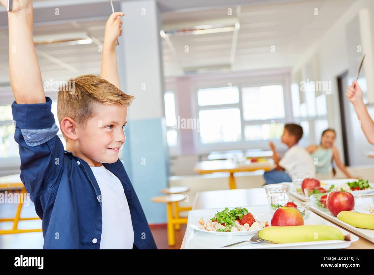 Lächelnder Junge mit erhobenen Armen, der während des Mittagessens in der Schulcafeteria am Tisch sitzt Stockfoto