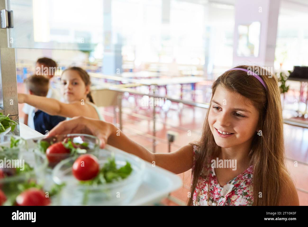Lächelnde Schülerin mit langen Haaren, die in der Mittagspause in der Schulcafeteria Gemüsesalat nimmt Stockfoto