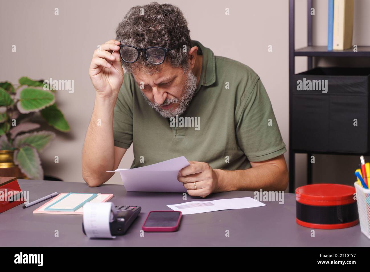 Ein fünfzigjähriger Mann mit Kurzsichtigkeit, mit grauem Bart verziert, hebt seine Brille, um einen Brief genau zu lesen, vielleicht eine Rechnung. Er ist lässig in einer Pole gekleidet Stockfoto