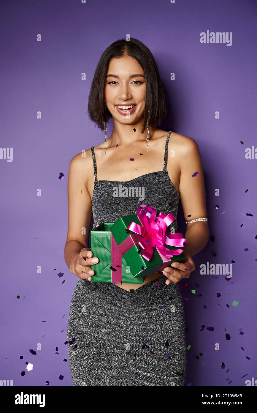 Glückliche asiatische Frau im Partykleid, die eingewickeltes Weihnachtsgeschenk in der Nähe von fallendem Konfetti auf lila hält Stockfoto