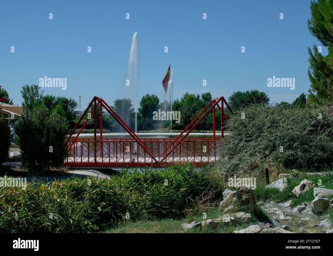 Rote Brücke über einen See mit zwei Springbrunnen in einem Park und spanischer Flagge, die im Hintergrund winkt Stockfoto
