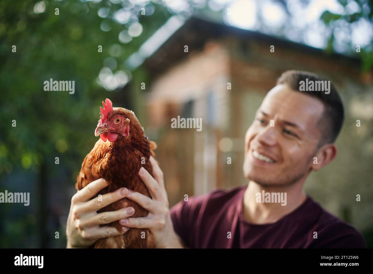 Porträt eines lächelnden Landwirts, der vor Hühnerstall steht und Hühner auf einem Bio-Bauernhof hält. Stockfoto
