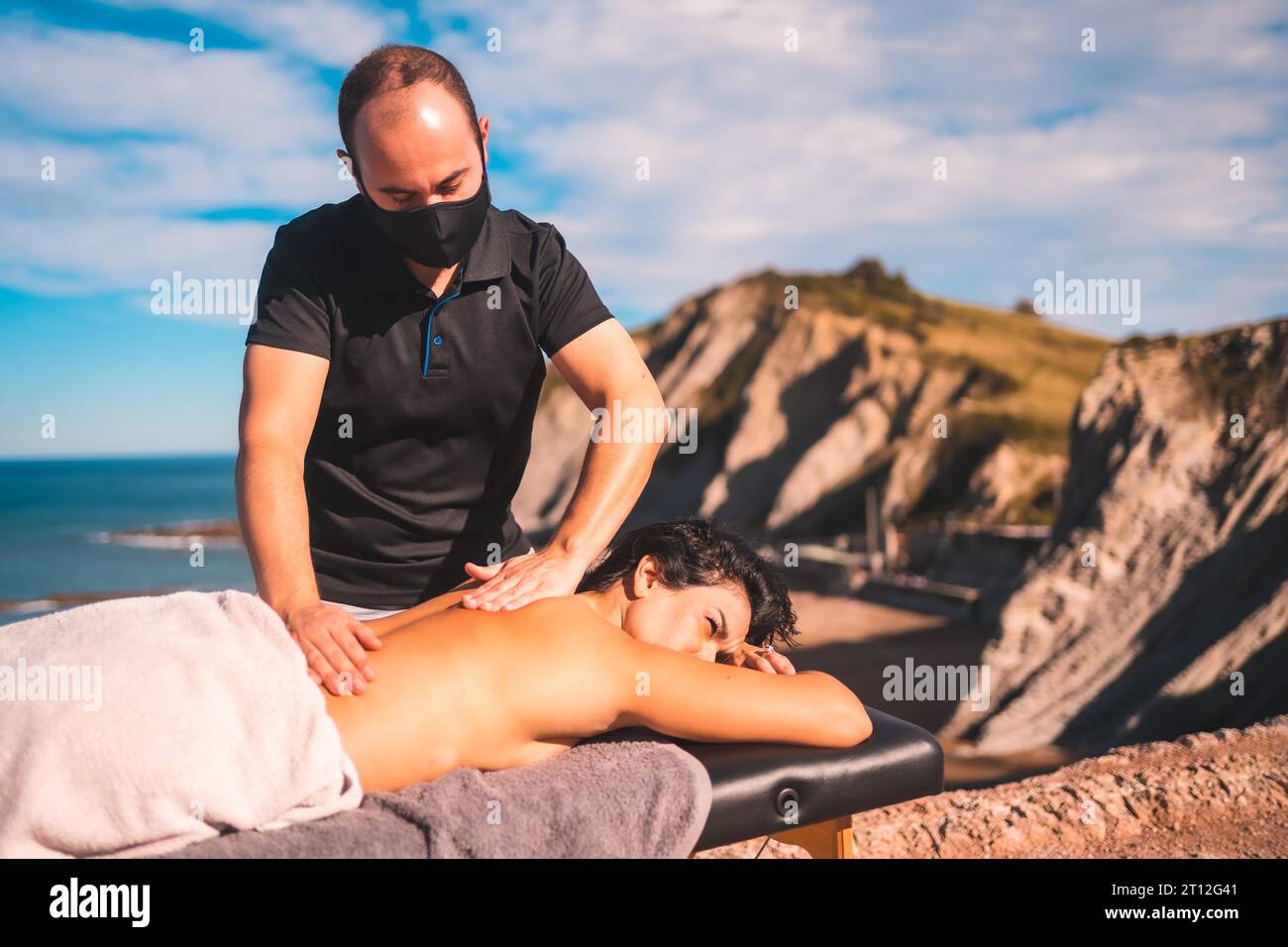 Entspanntes Mädchen bei der Massage an der Küste nahe dem Meer, Masseurin mit Gesichtsmaske in der Coronavirus-Pandemie Stockfoto