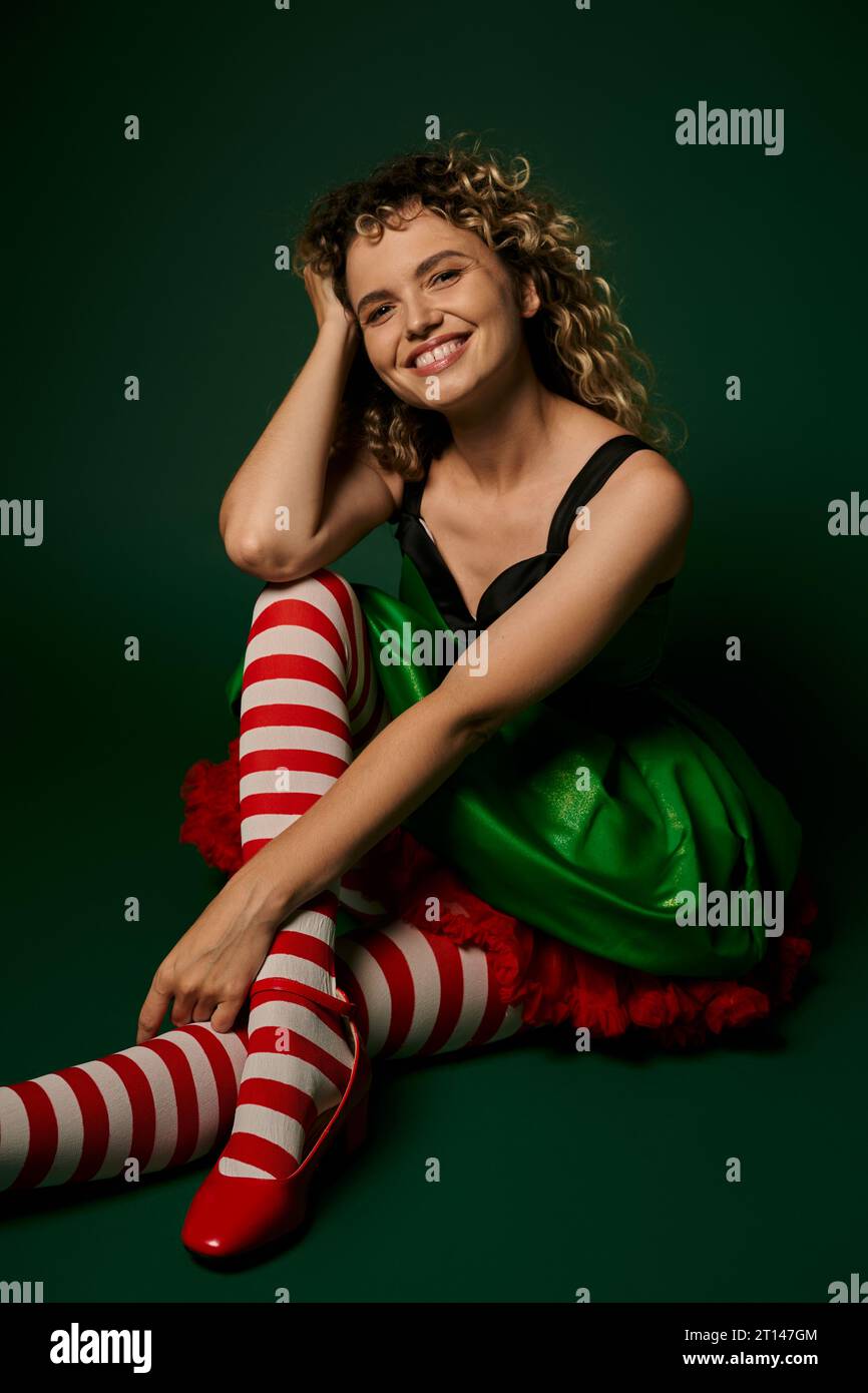 Fröhliche Frau im Silvester-Elfenkostüm und gestreiften Strümpfen lächelt vor grüner Kulisse in die Kamera Stockfoto
