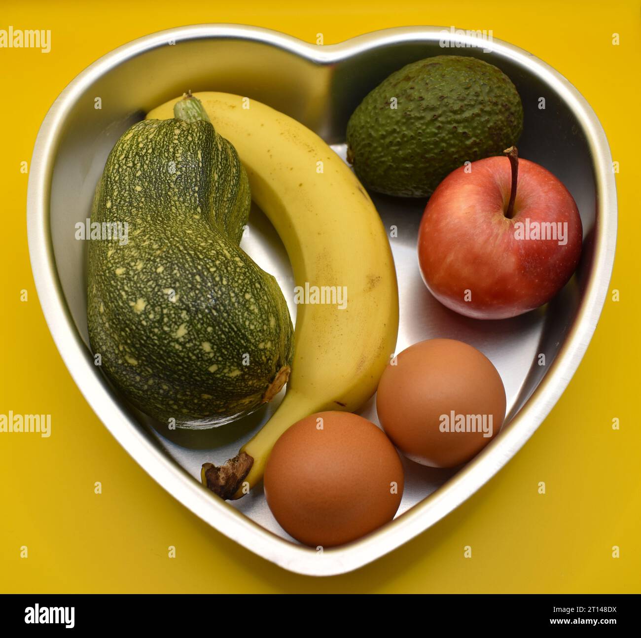 Ein gesundes Herz, eine Auswahl an Obst-, Gemüse- und Milchprodukten in einer herzförmigen Backform vor gelbem Hintergrund. Stockfoto