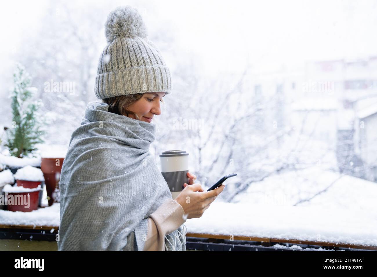 Eine Frau in einem warmen Hut und einer Decke hält das Smartphone in den Händen. Winterlandschaft mit Schneefall im Hintergrund. Stockfoto