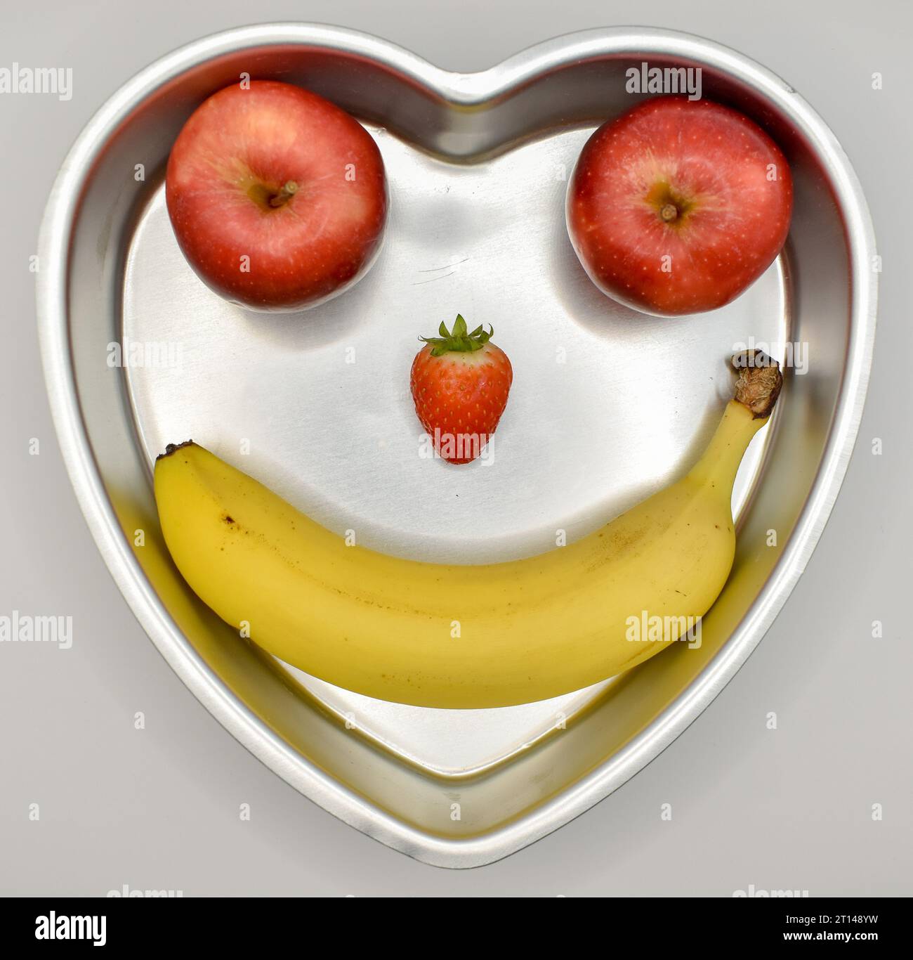 Essen Sie gesund, eine herzförmige Dose mit zwei Äpfeln, einer Erdbeere und einer Banane, die ein lächelndes Gesicht bilden. Stockfoto