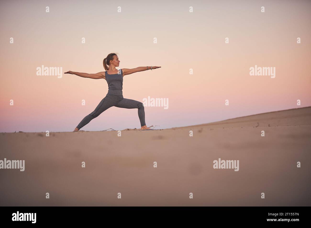 Schöne junge Frau praktiziert Yoga Asana Virabhadrasana 2 - Kriegerposition 2 in der Wüste bei Sonnenuntergang. Stockfoto