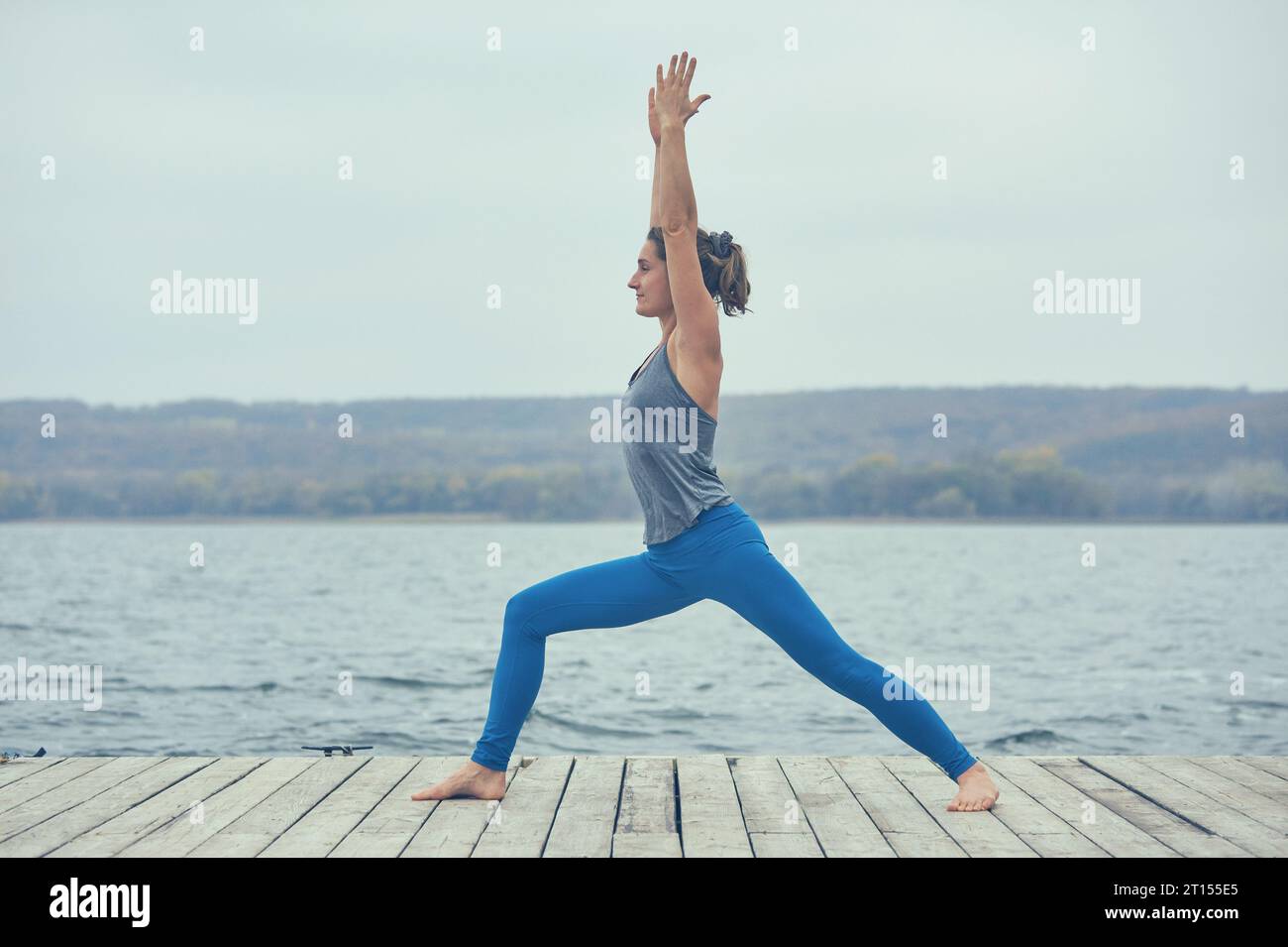 Schöne junge Frau praktiziert Yoga Asana Virabhadrasana 1 - Kriegerposition 1 auf dem Holzdeck in der Nähe des Sees. Stockfoto