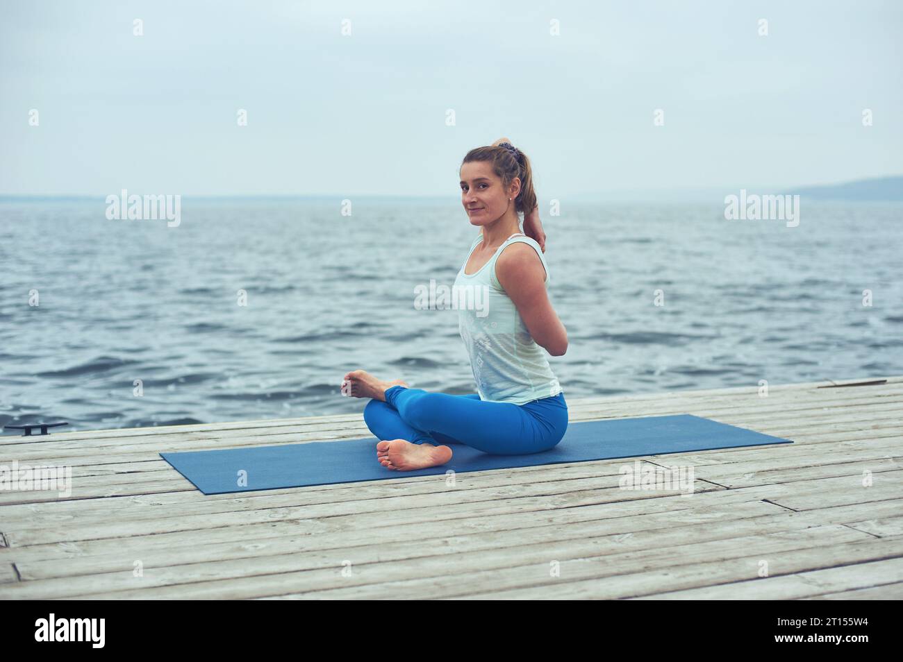 Schöne junge Frau praktiziert Yoga Asana Gomukhasana - Kuhgesicht Pose auf der Holzterrasse in der Nähe des Sees. Stockfoto