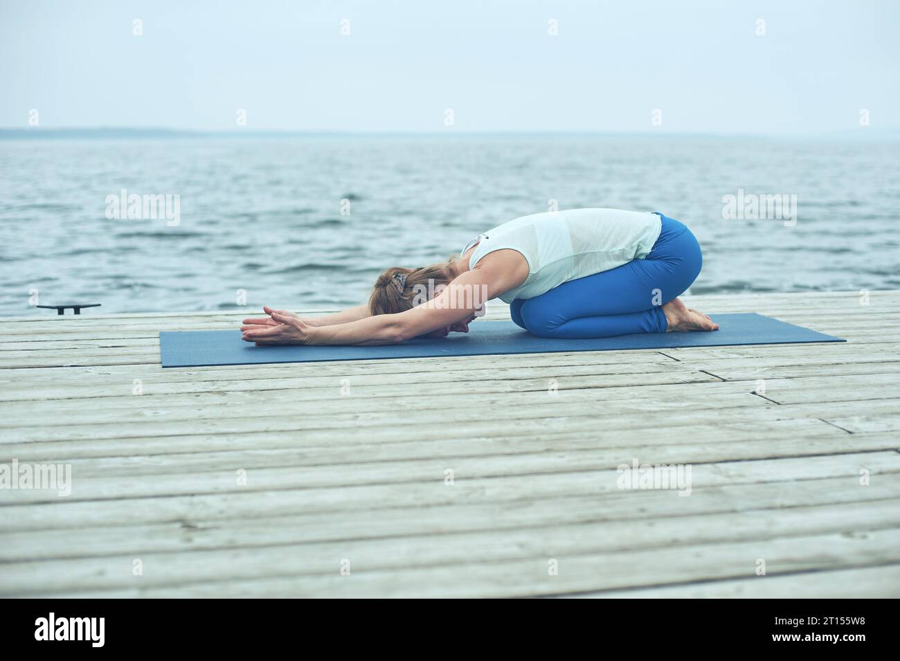 Schöne junge Frau praktiziert Yoga Asana Gomukhasana - Kuhgesicht Pose auf der Holzterrasse in der Nähe des Sees. Stockfoto