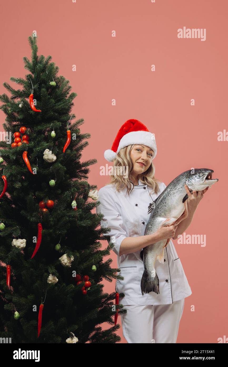 Das Konzept des Kochens und der Weihnachtsferien. Eine schöne Köchin, die einen großen Fisch in der Hand hält, vor dem Hintergrund eines Weihnachtsbaumes. Stockfoto