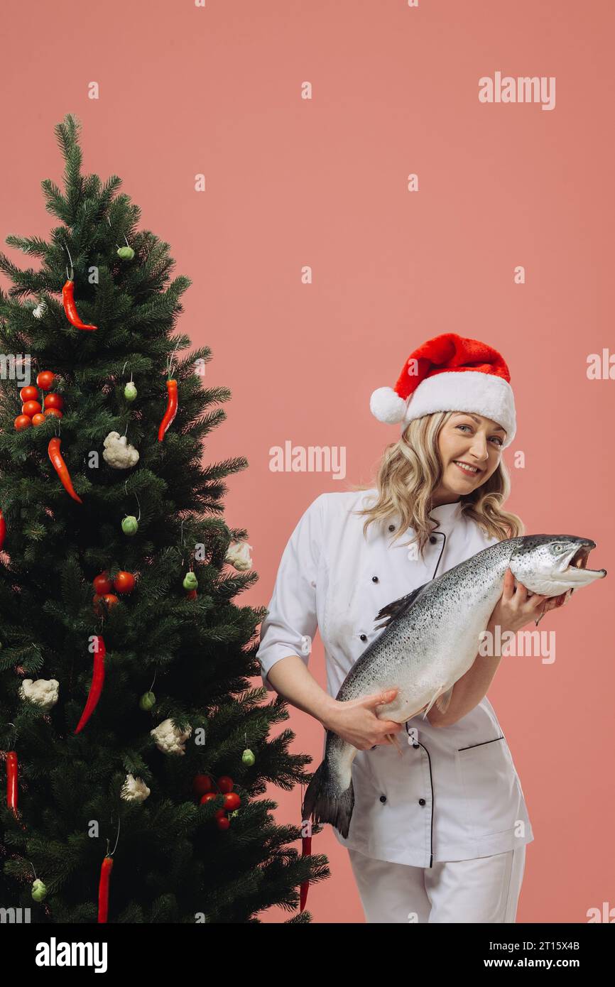 Das Konzept des Kochens und der Weihnachtsferien. Eine schöne Köchin, die einen großen Fisch in der Hand hält, vor dem Hintergrund eines Weihnachtsbaumes. Stockfoto